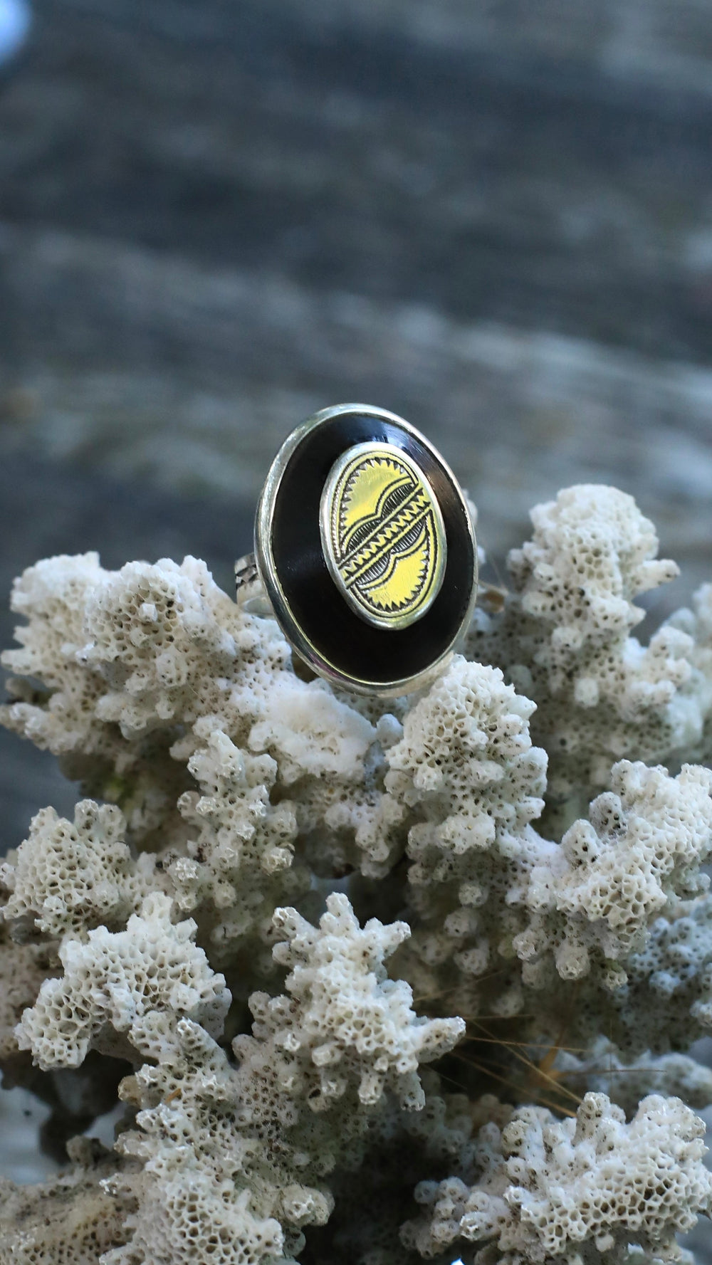 A silver-toned oval ring with a black enamel face and a yellow-and-black central emblem, displayed on white coral against a blurred gray background.