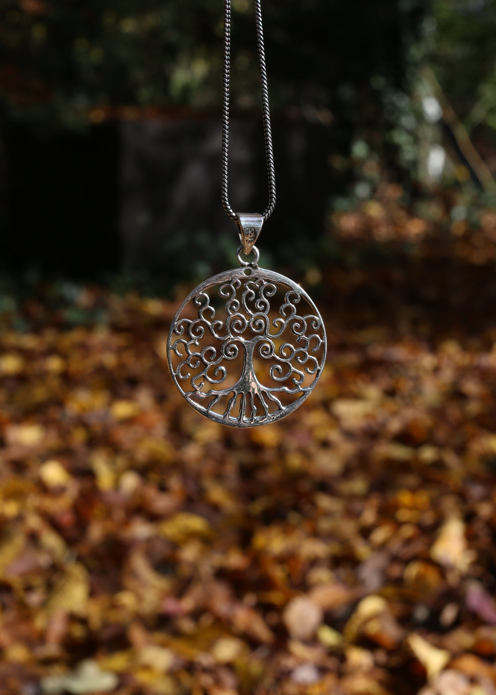 Silver tree of life pendant hanging against a natural background with autumn leaves